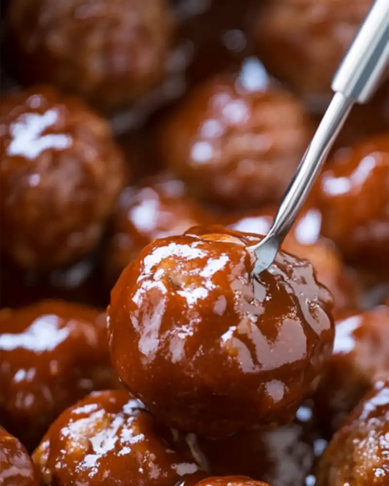 A close-up of glazed BBQ meatballs in a crockpot covered in a shiny, brown sauce, with a fork holding up one meatball above the rest.