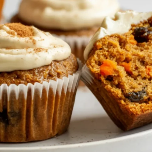 A moist gluten-free carrot muffins made with almond flour, topped with cream cheese frosting, sits on a white plate showing its fluffy texture and visible carrot pieces.
